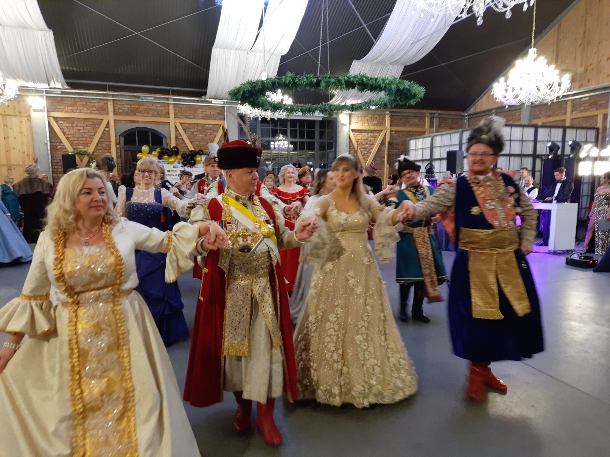 Ubrani ludzie tańczą podczas balu w strojach historycznych. Widać mężczyzn i kobiet w bogatych, barwnych ubiórach z koronkami i złotymi dodatkami.