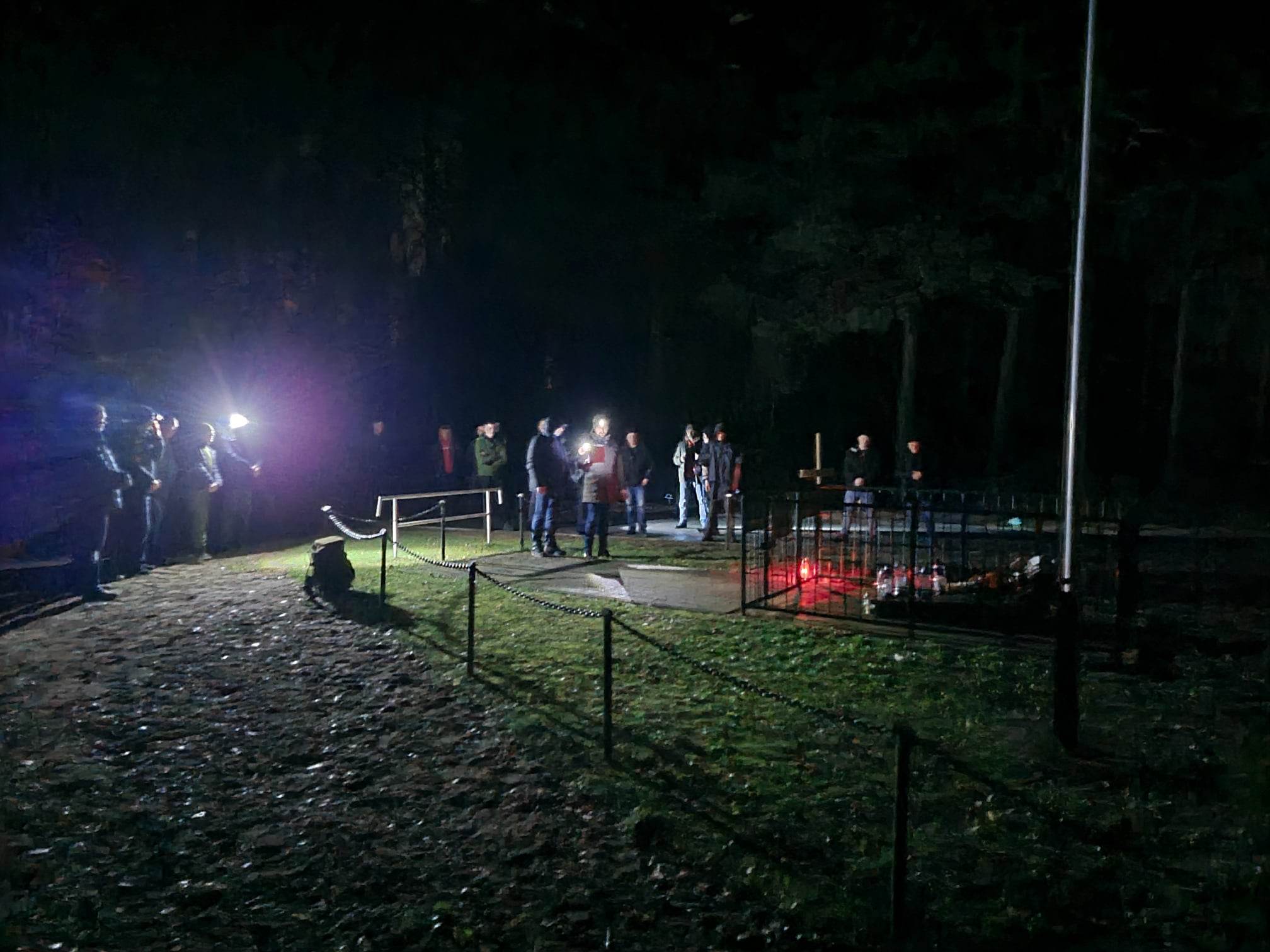 Na ciemnym tle widać grupę osób stojących na trawiastym terenie. W oddali widoczna jest konstrukcja ogrodzeniowa z metalowymi słupami i siatką. Kilka źródeł światła oświetla scenę.