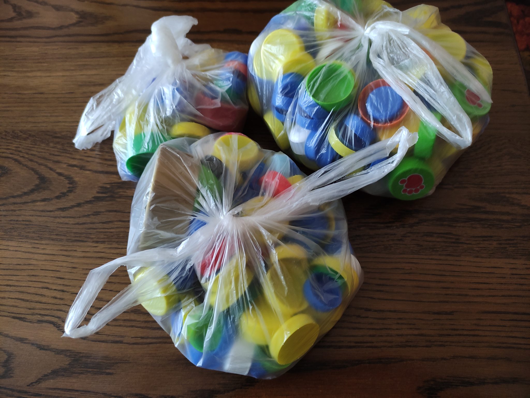 Zdjęcie trzech worków wypełnionych nakrętkami plastikowymi. Nakrętki są w różnych kolorach, a worki związane sznurkami. Na drewnianej powierzchni tło.