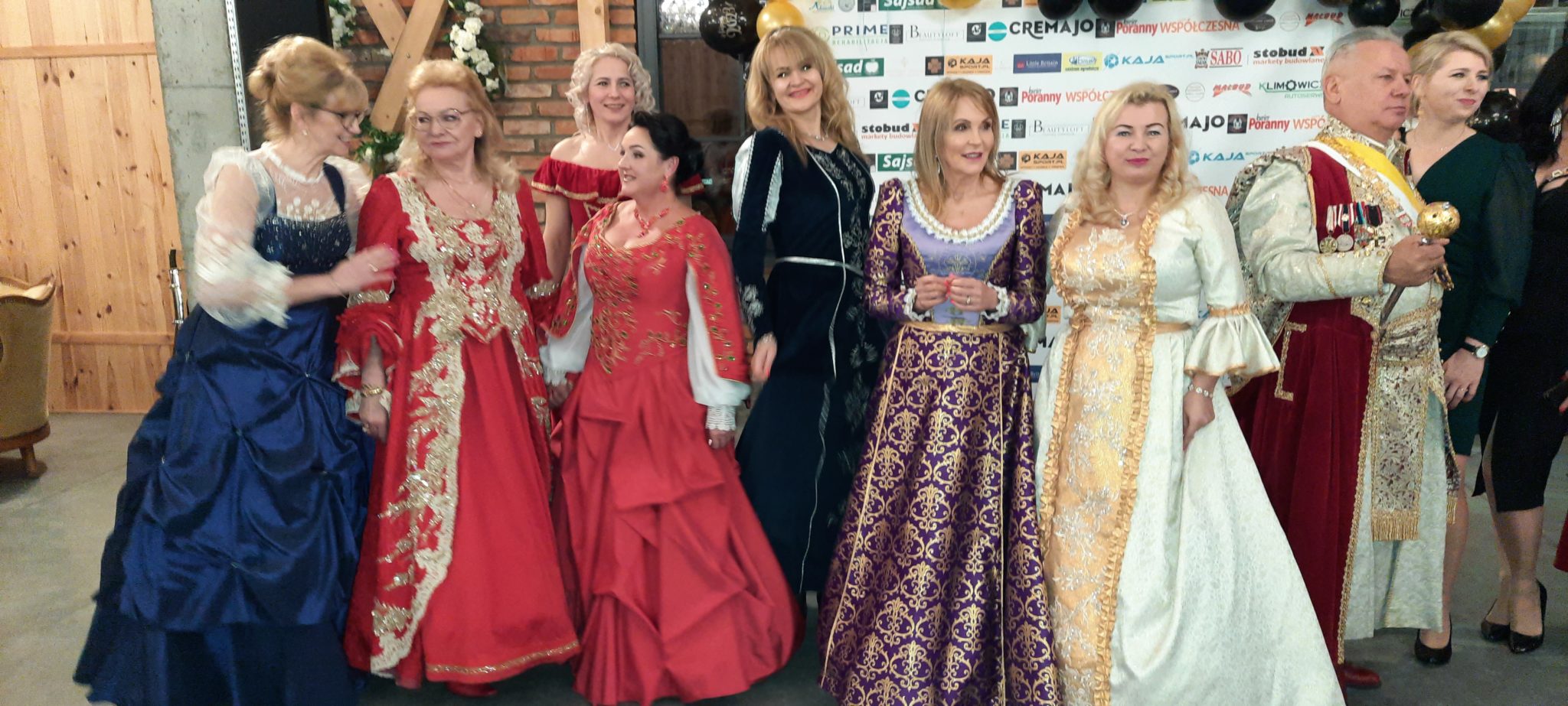 Grupa osób w strojach historycznych bierze udział w balu charytatywnym. Widać kobietę w czarnym платье, kobietę w czerwonym платье z złotymi zdobieniami oraz kilka innych osób w podobnych stylizacjach.