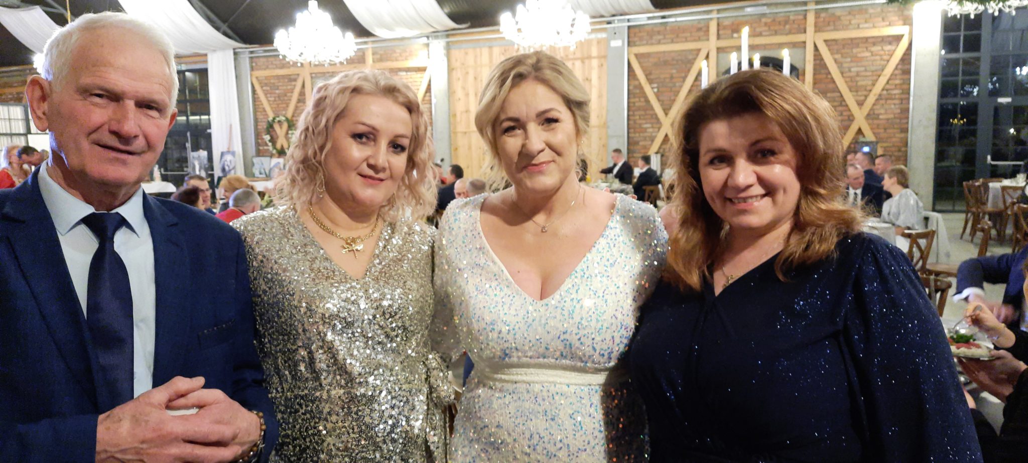 Grupa osób w eleganckich strojach na balu charytatywnym. Widać mężczyznę w marynarce i krawacie oraz trzy kobiety w błyszczących sukniach.