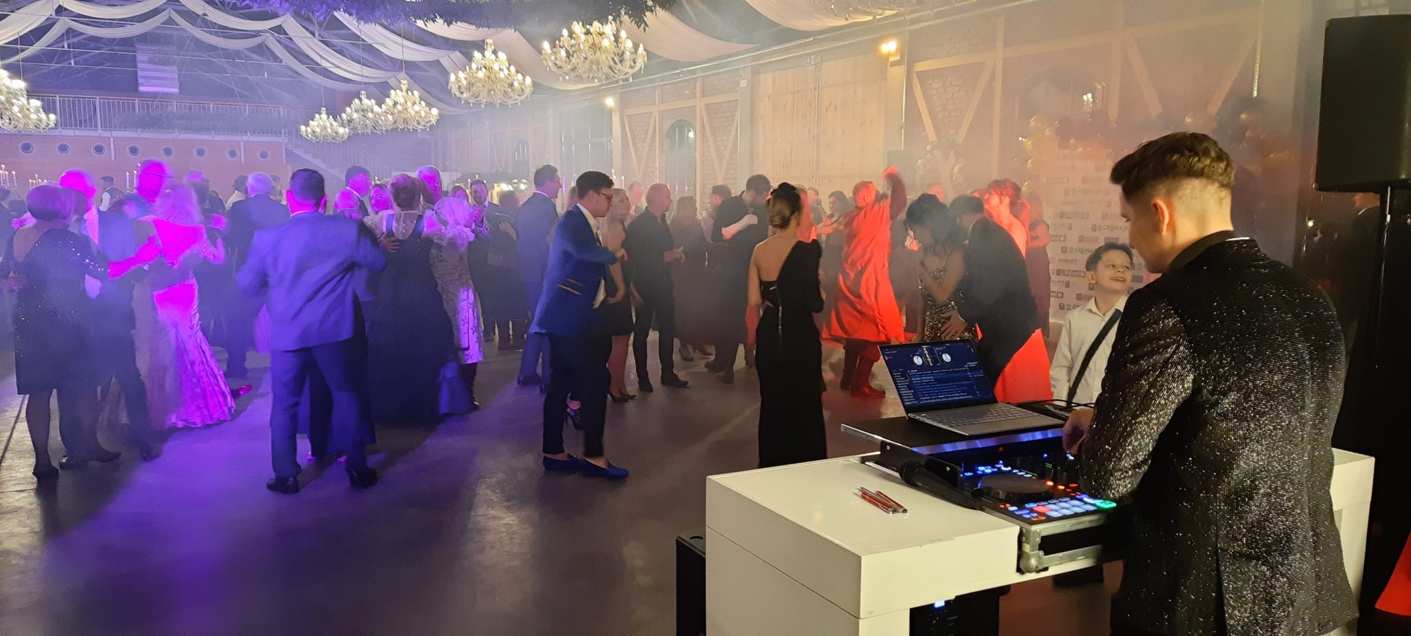 W sali balowej widać ludzi tańczących i osobę obsługującą sprzęt muzyczny. Ściany są oświetlone kolorowymi światłami, co tworzy atmosferę wydarzenia.