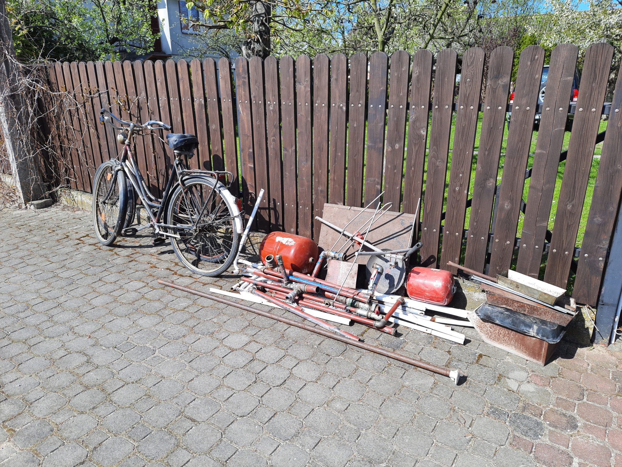 Na chodniku znajduje się rower, narzędzia i części zderzaka, stojące przy drewnianym płocie.