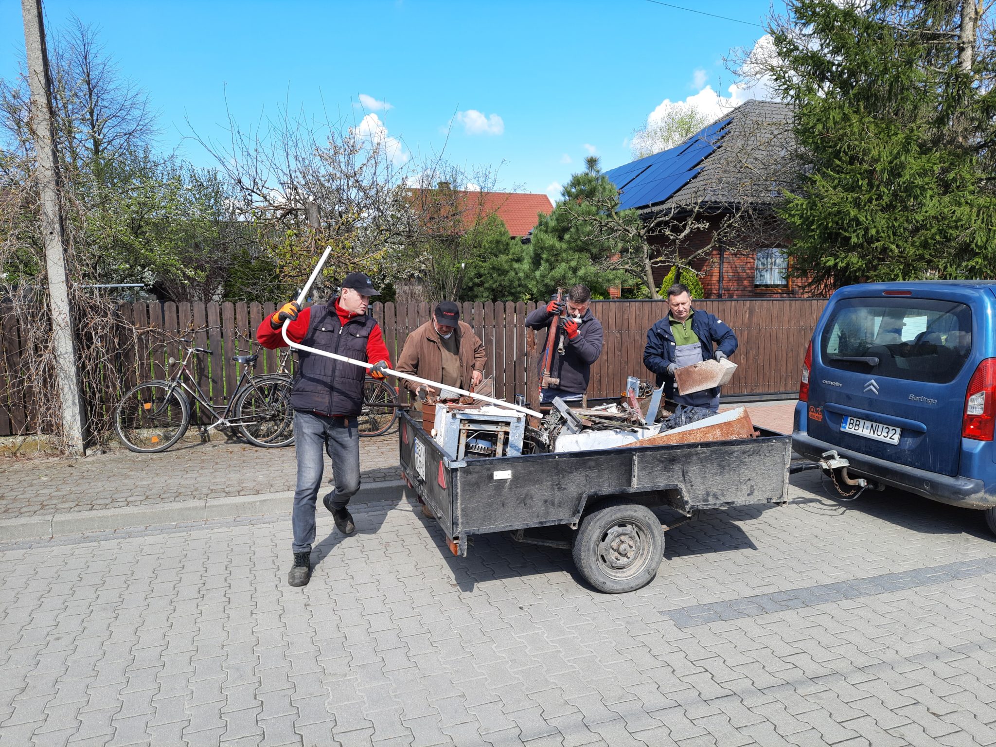 Zdjęcie przedstawia zbiórkę złomu w Brańsku. Cztery osoby transportują na przyczepce różne metalowe elementy i sprzęt. W tle zielony płot, drzewa oraz niebo.
