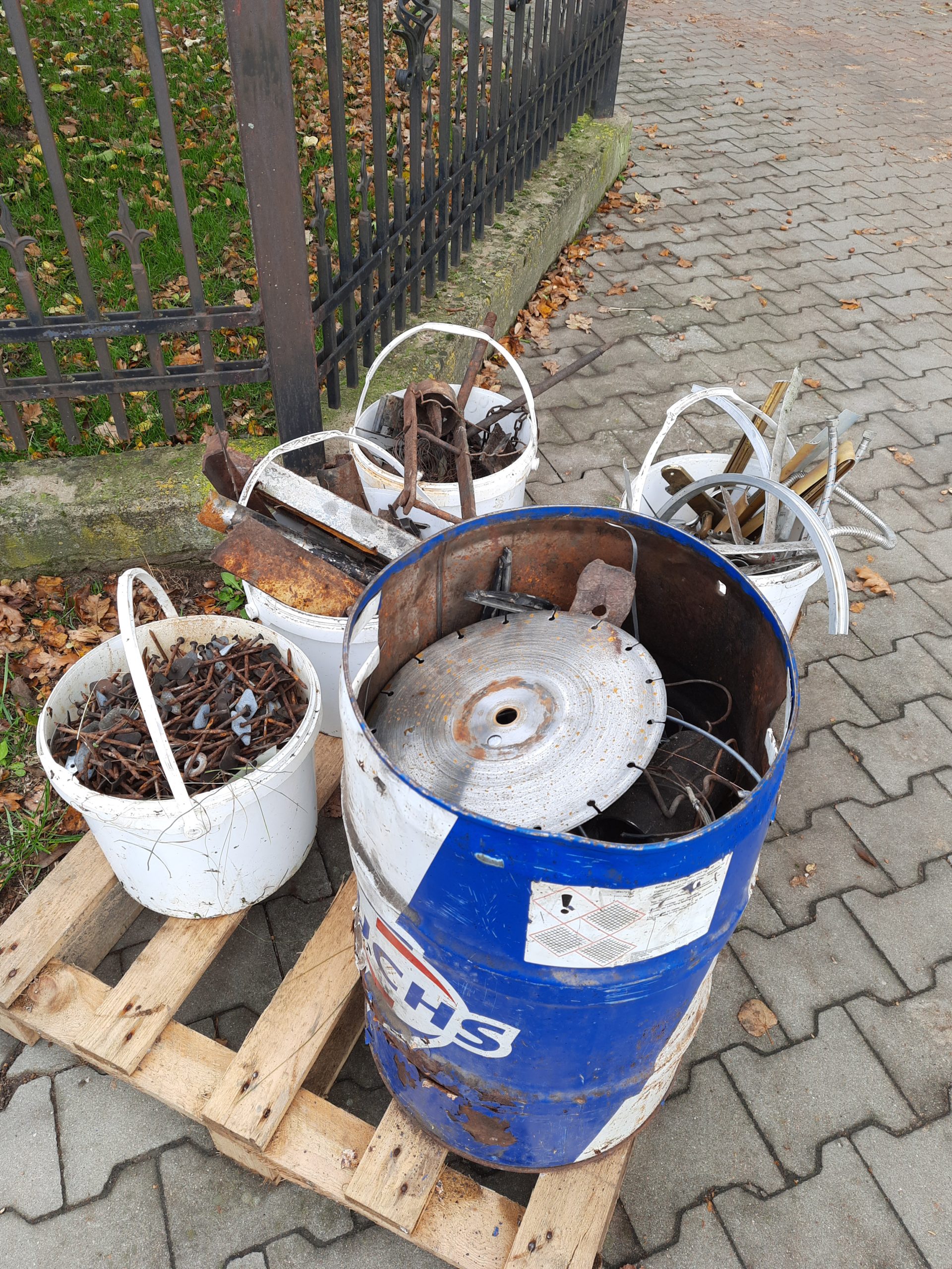 Zdjęcie przedstawia trzy białe kosze i metalową beczkę z elementem przypominającym koło, które leżą na drewnianej palecie na chodniku. W tle znajduje się płot i liście opadające z drzew.