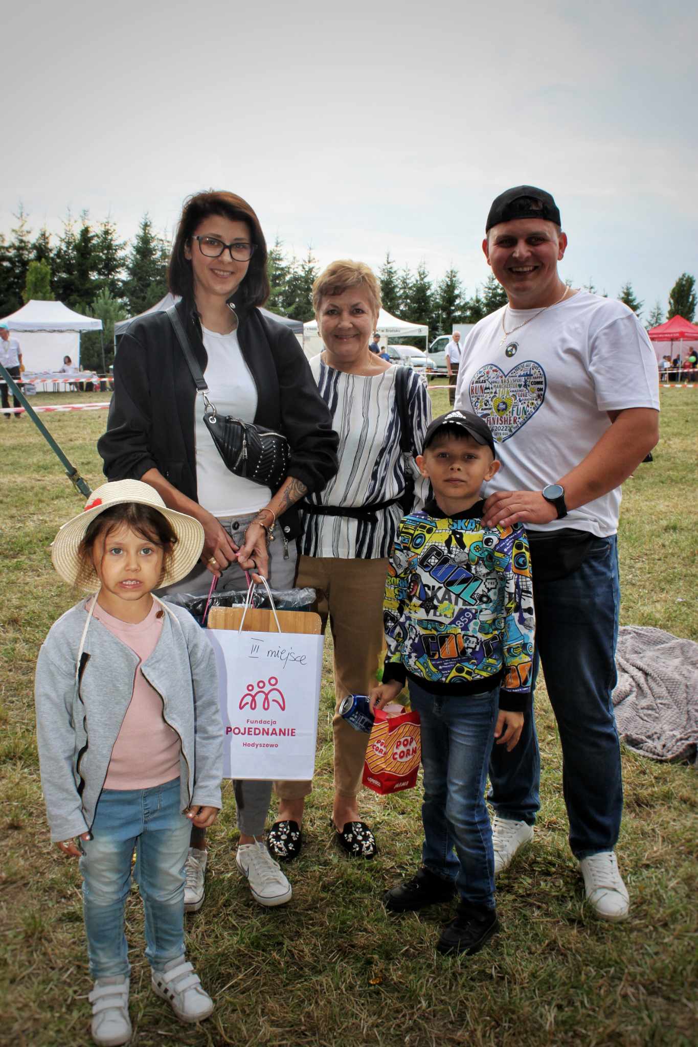 Rodzina na pikniku. Dziecko w kapeluszu trzyma torbę, a reszta rodziny stoi obok. W tle krajobraz z drzewami i budynkami. Opisuje to scenę z wydarzenia „W Dzień Rodziny”.