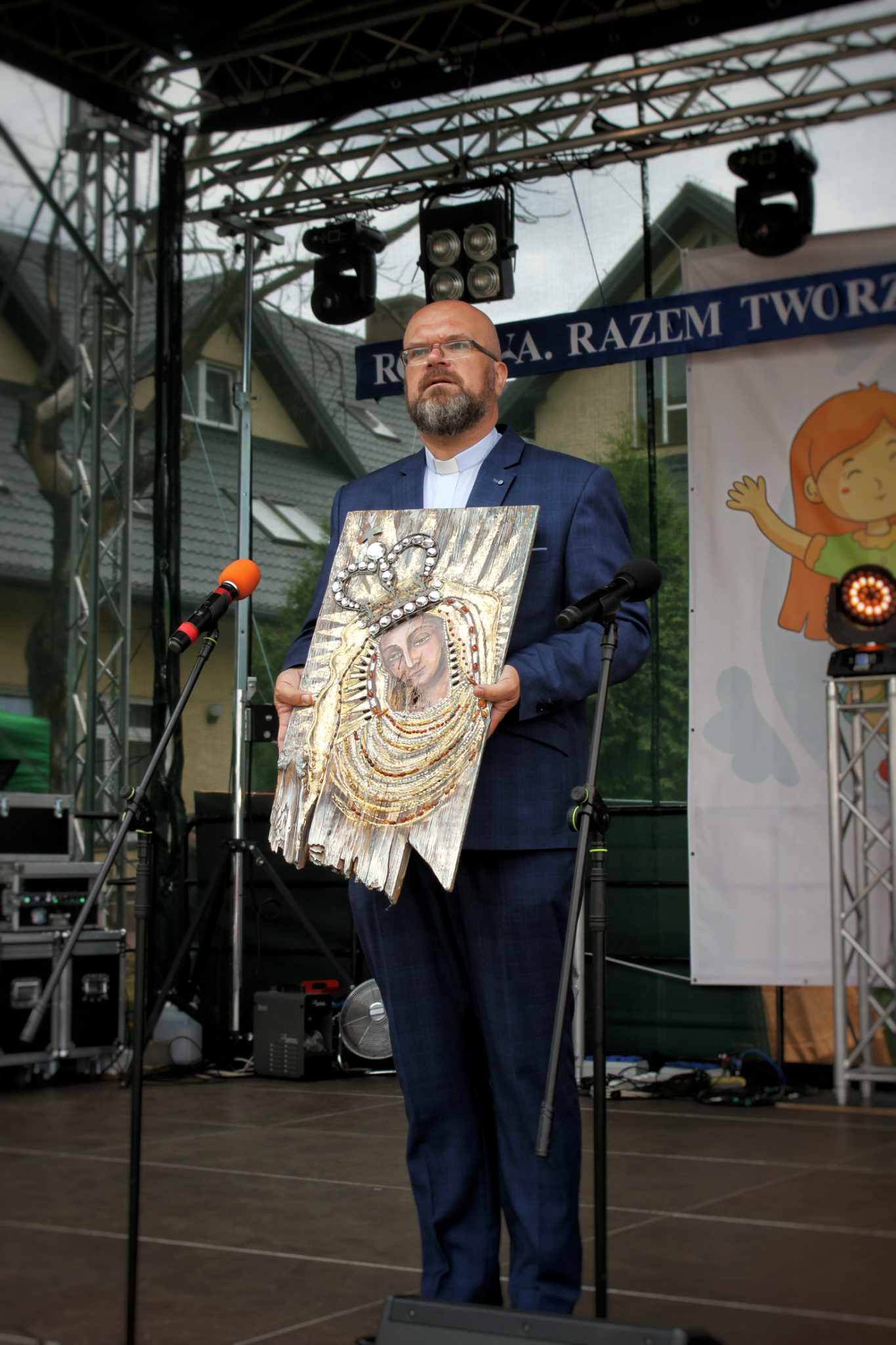 Kapłan trzyma ozdobny wizerunek Matki Bożej na scenie podczas wydarzenia w Hodyszewie.