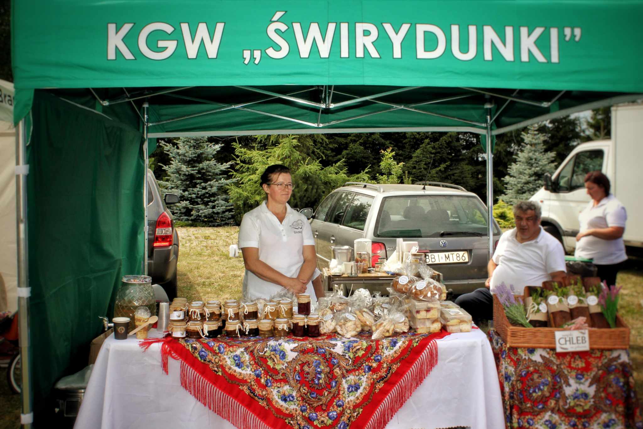 Na stoisku targowym KGW Świrydunki wystawione są produkty spożywcze, w tym dżemy i miody. W tle zielona markiza.