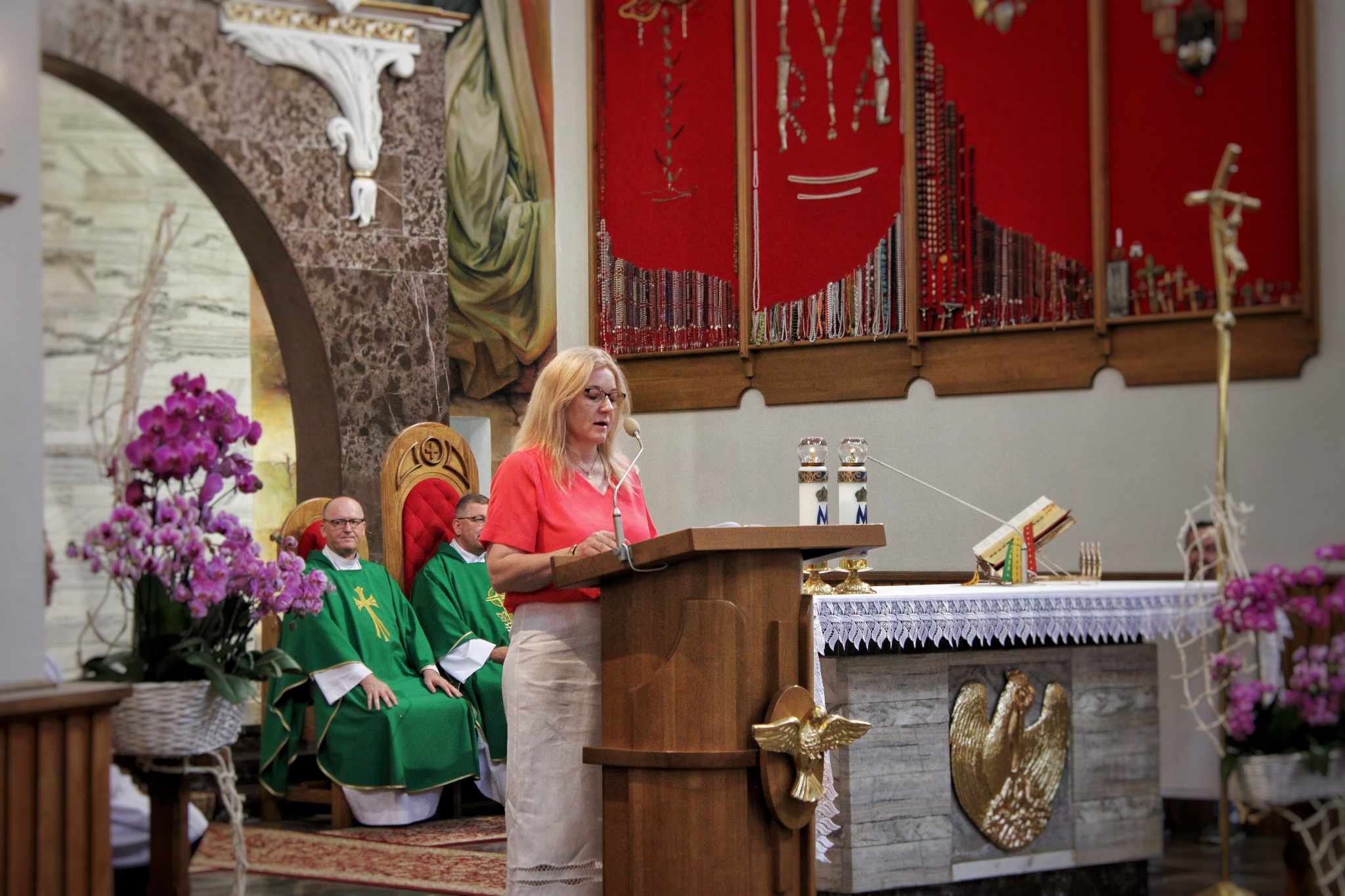Wnętrze kościoła, podczas uroczystości Dnia Rodziny. Kobieta w czerwonym płaszczu stoi przy ambonie, otoczona przez mężczyzn w zielonych szatach. W tle znajdują się obrazy i kwiaty.