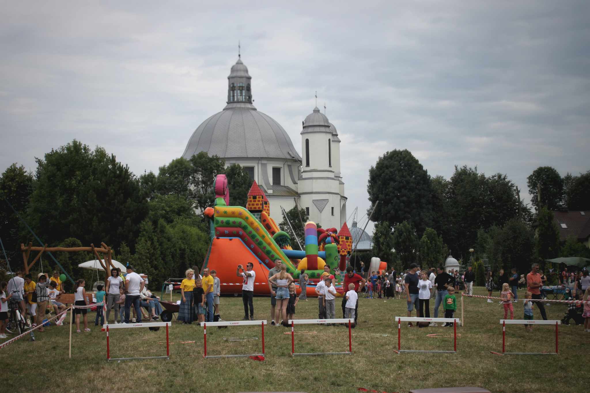Na placu festynowym znajduje się zjeżdżalnia i inne atrakcje dla dzieci. W tle widoczna jest duża, biała budowla o kopułowym dachu. Uczestnicy imprezy bawią się na placu.