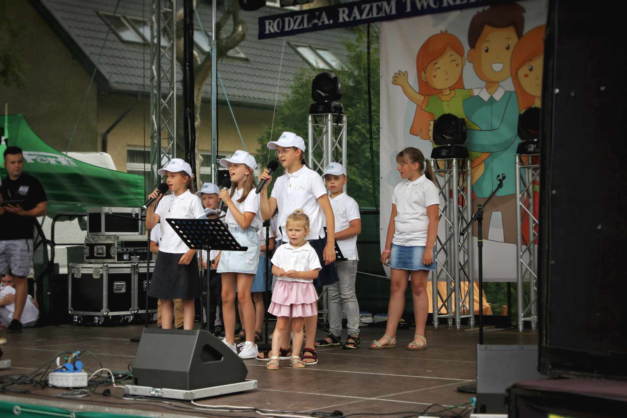 Na scenie grupa dzieci w białych koszulach z kapeluszami prezentuje się podczas wydarzenia. W tle plakat z napisem 'BZOŁ-A, RAZEM TWÓJ'.