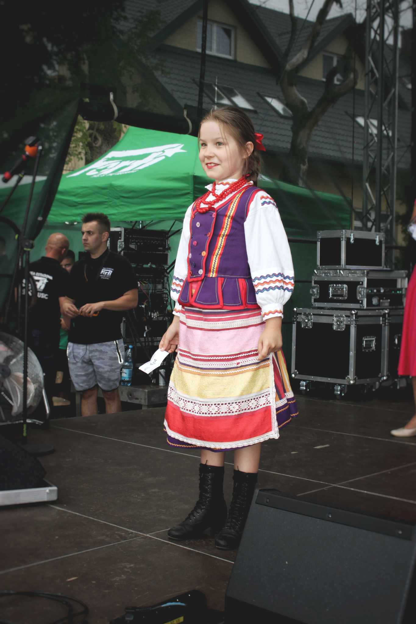 Na scenie stoi młoda osoba ubrana w tradycyjny strój ludowy. Ma na sobie białą koszulkę, bordową kamizelkę i spódnicę z kolorowymi paskami. Na nogach ma czarne buty. W tle widać sprzęt muzyczny oraz ludzi.