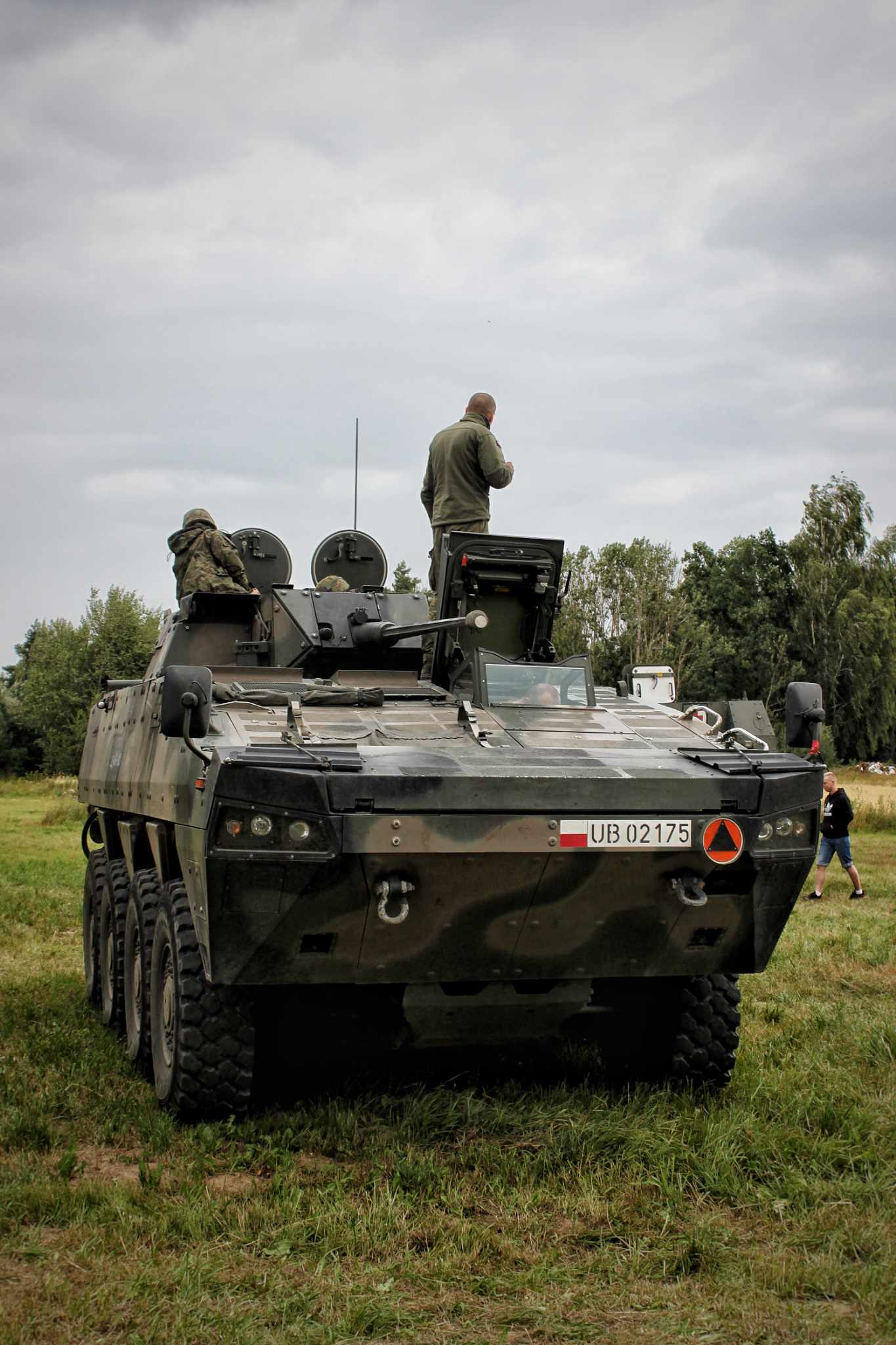 Pancerny wóz rozpoznawczy z podwoziem na sześciu kołach, pomalowany w kamuflaż, stoi na trawiastym terenie. Wewnątrz i na dachu znajdują się osoby w uniformach wojskowych. Na tle zielone drzewa.