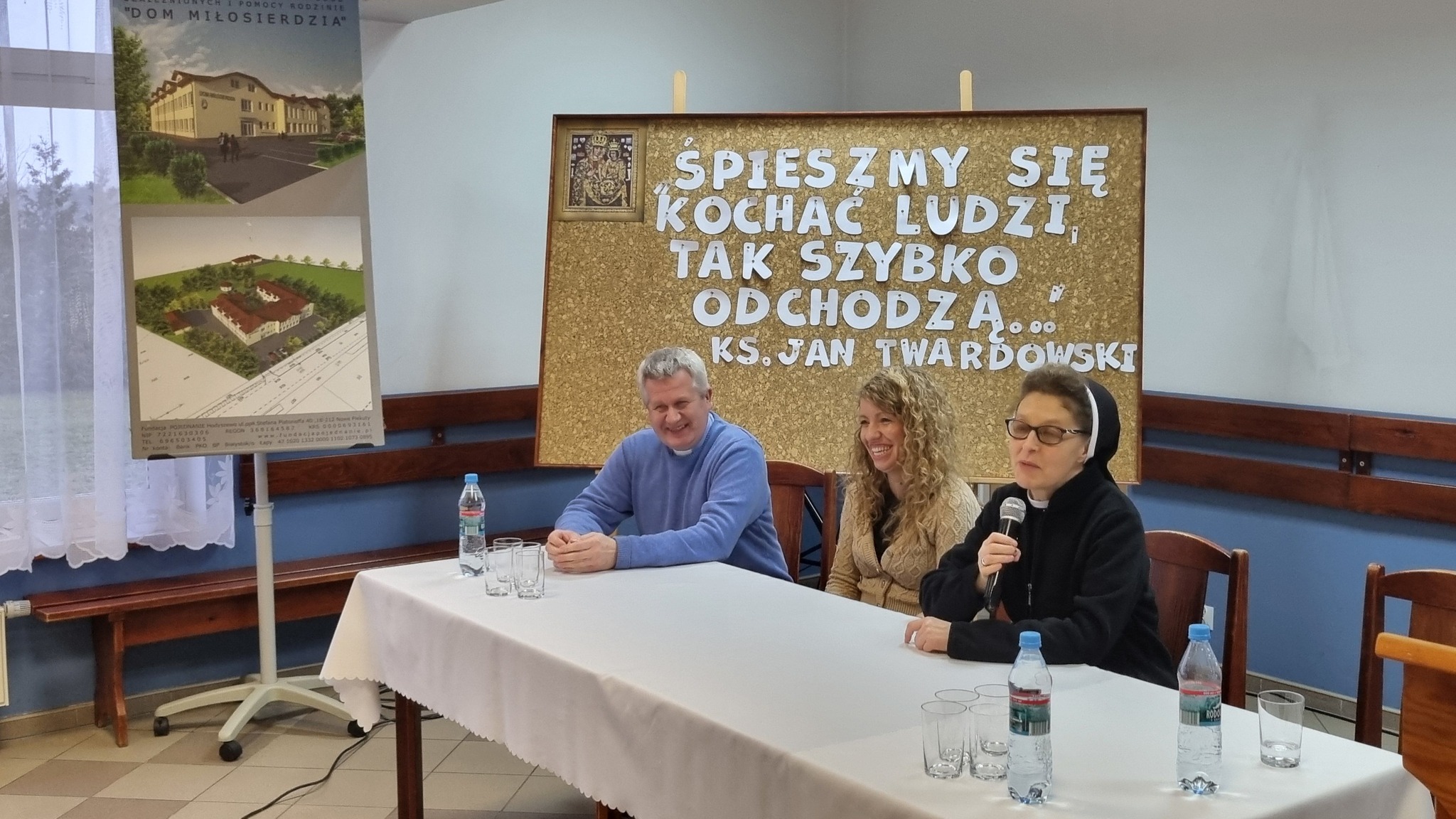 Na stole siedzą trzej ludzie podczas spotkania. Na ścianie znajduje się plakat z napisem "Śpieszmy się, kochać ludzi, tak szybko odchodzą".