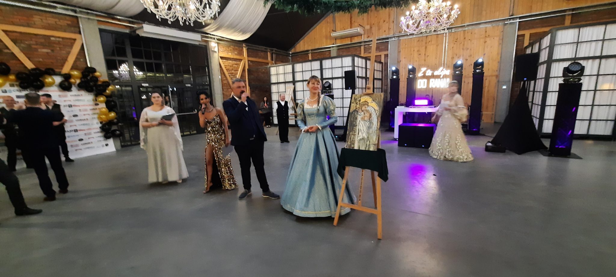 Grupa osób na balu charytatywnym wewnątrz budynku z dekoracjami świetlnymi i muzycznym sprzętem.