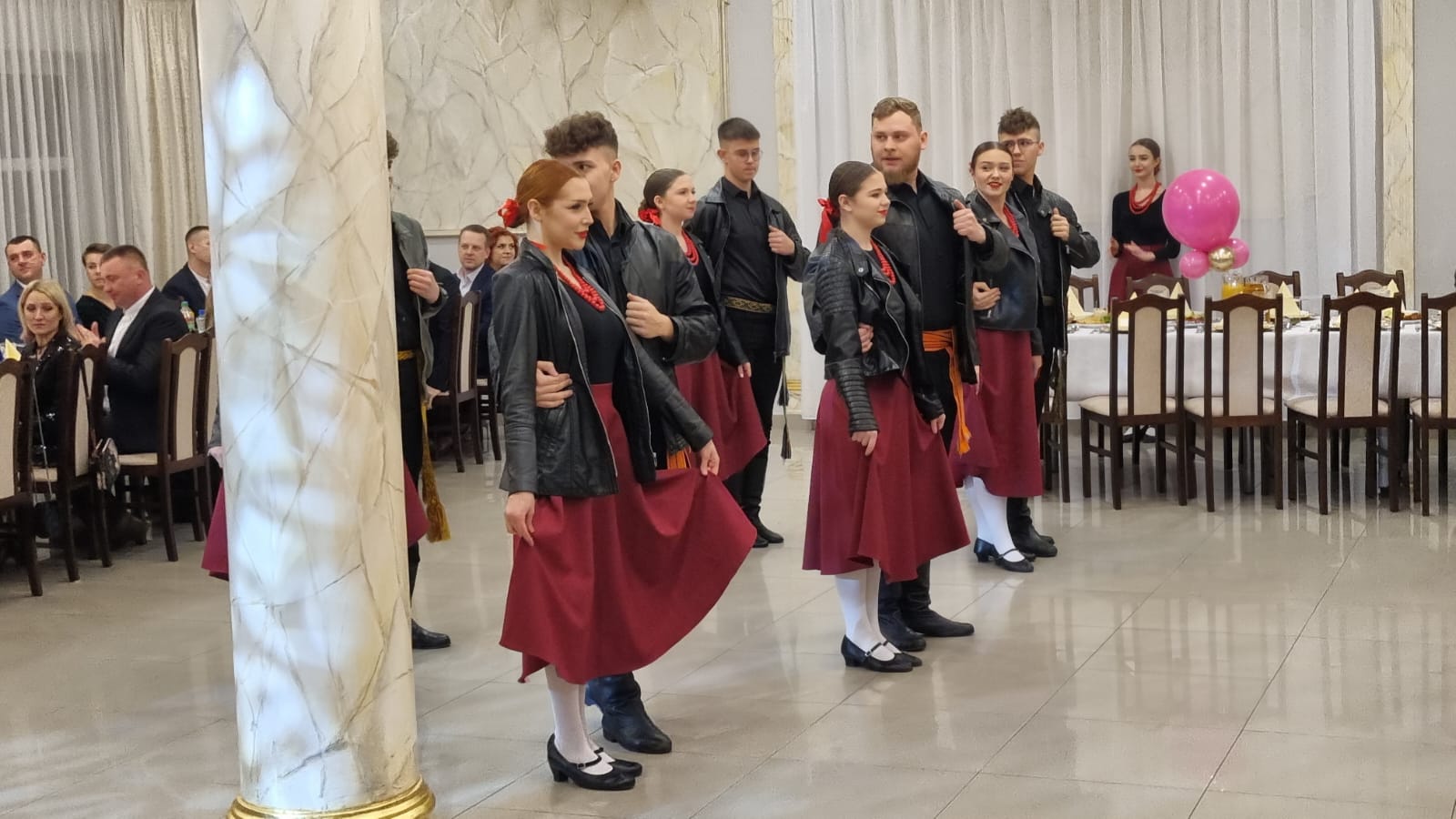 Grupa tańczących osób w strojach tradycyjnych na balu charytatywnym. Widać mężczyznę i kobiety w czerwonych spodniach, czarnych kurtkach oraz białe koszule. Goście ubrani są w formalne stroje. Odbicie światła na kolumnie z marmurowym wzorem.