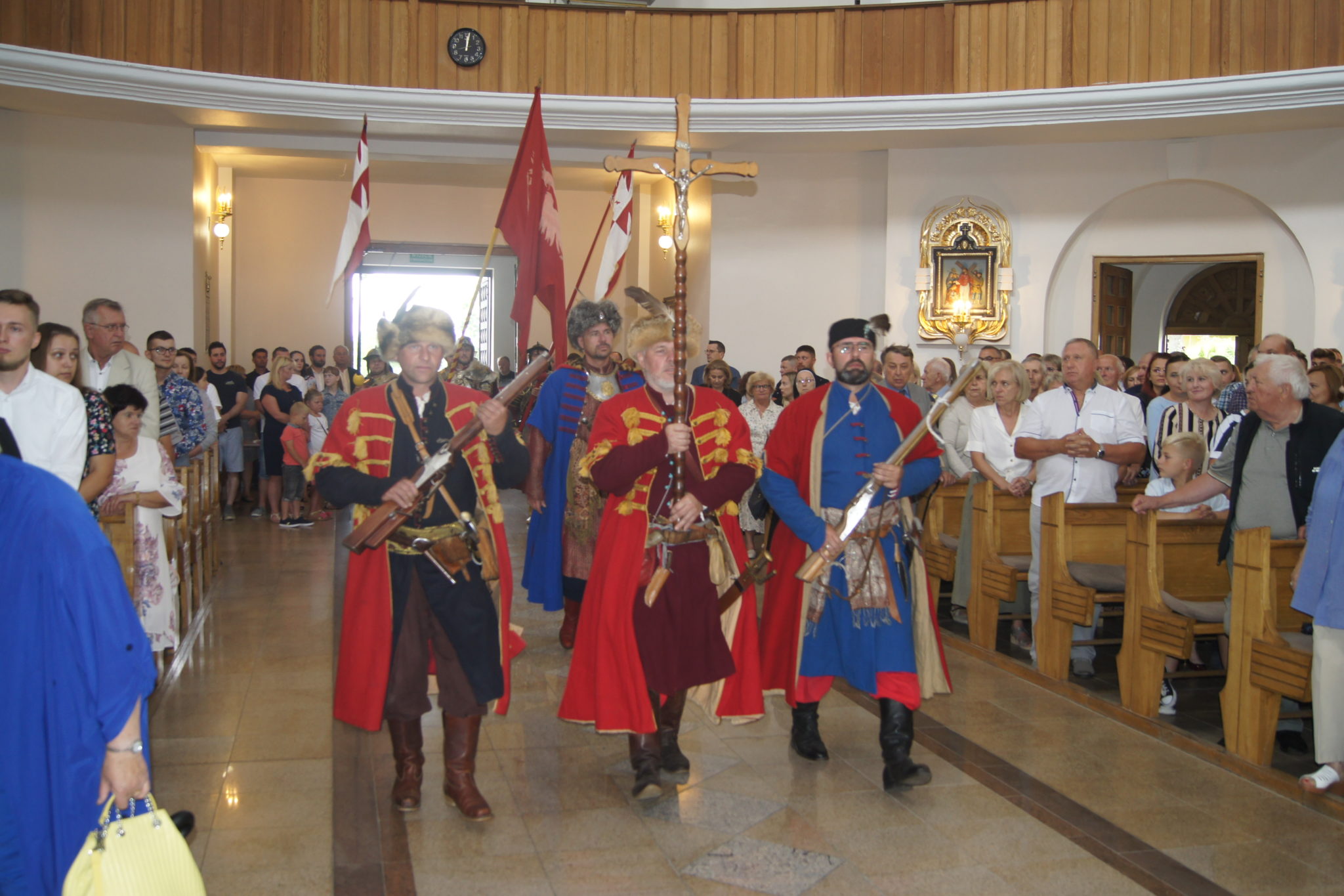 Grupa osób w strojach ceremonialnych przechodzi przez kościelny podłogę. Wszyscy są ubrani na czarno, a osoby w strojach mają czerwone i niebieskie elementy. W tle widoczna publiczność siedząca na ławkach.