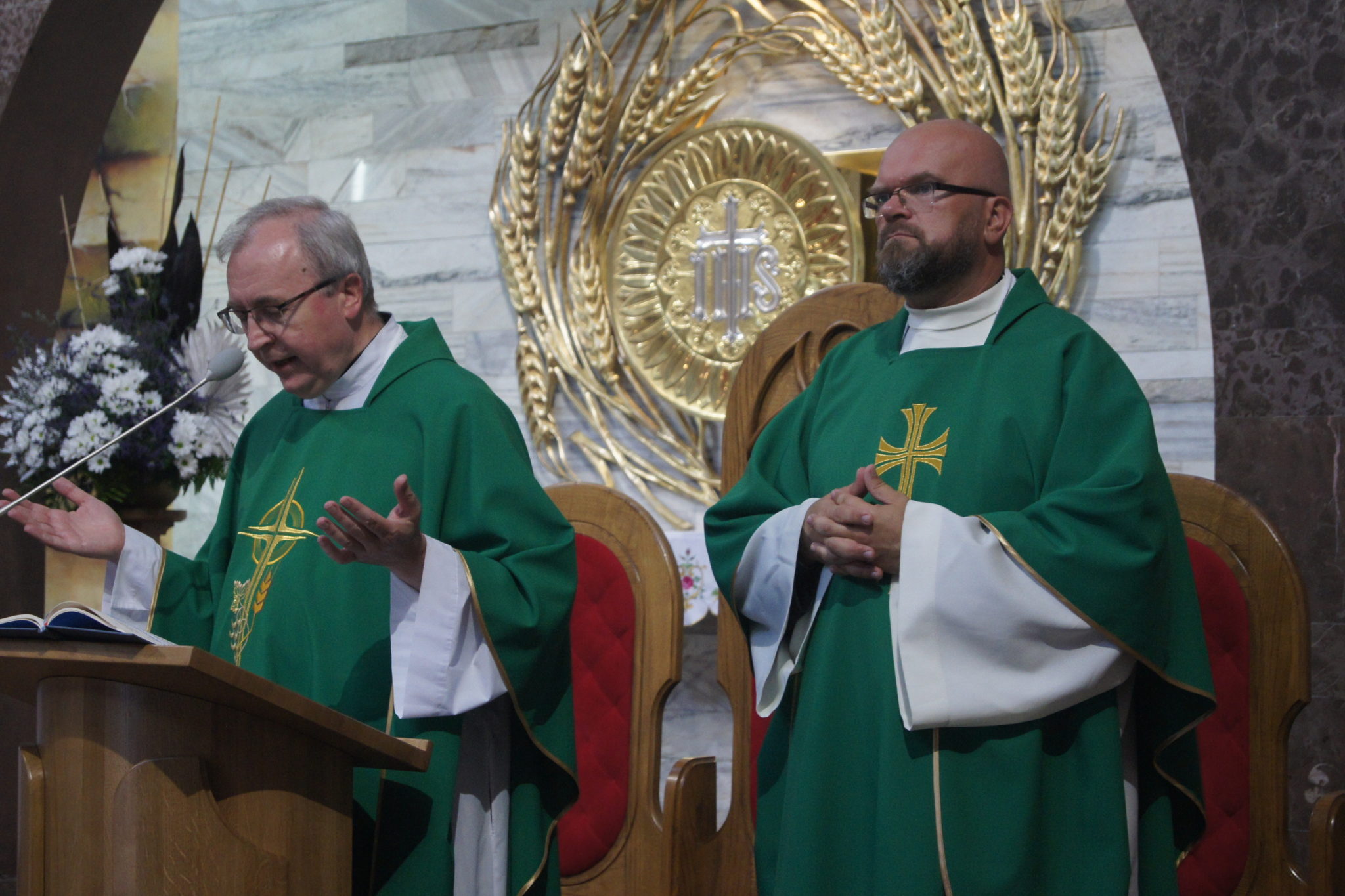 Na zdjęciu widoczni dwoje mężczyzn ubranych w zielone szaty z białymi rękawami i krzyżami. Jeden z mężczyzn stoi za ambony, trzymając księgę, a drugi siedzi na krześle. W tle znajduje się złoty obraz z symbolem wieńca i krzyżem.