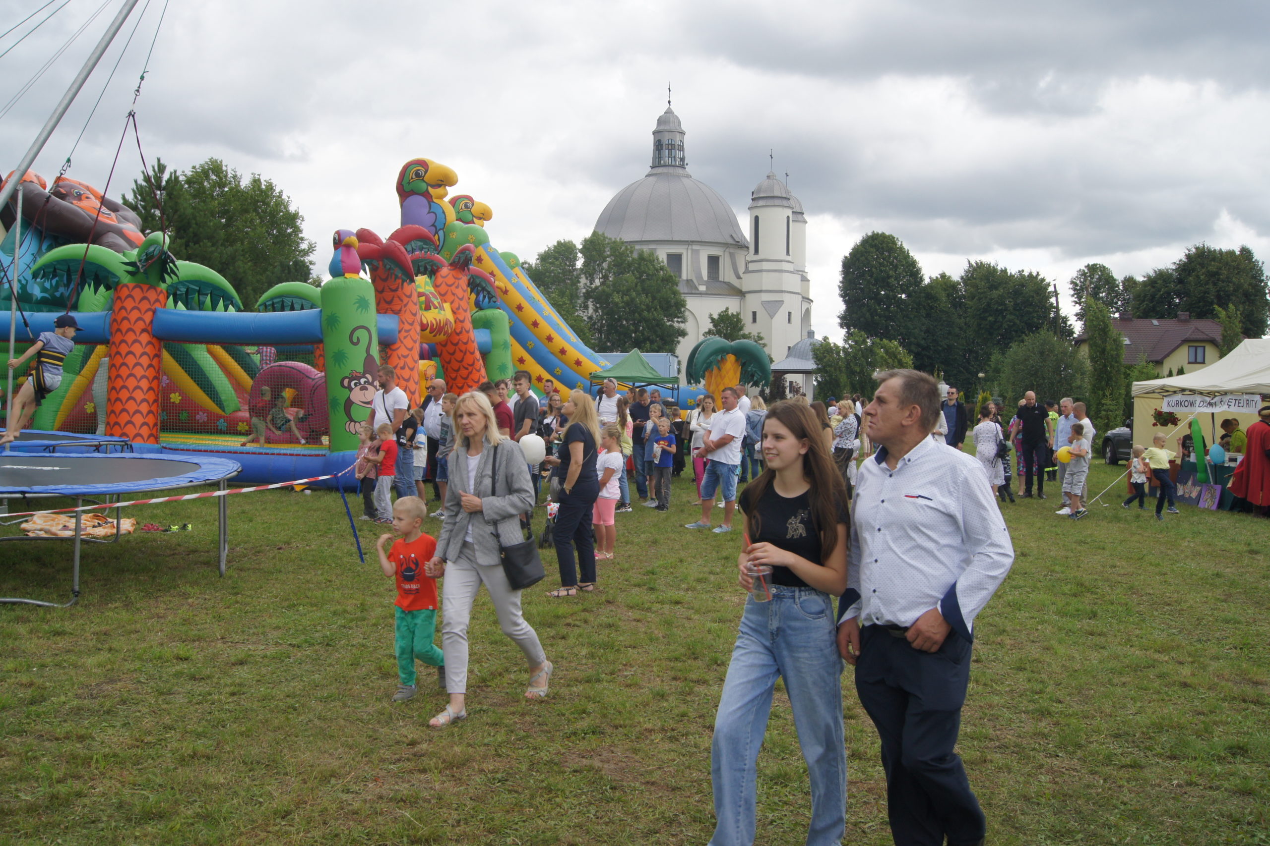 Na placu odbywa się wydarzenie, na którym obecni są ludzie i znajduje się dmuchany domek z animacjami oraz kościół. Organizatorem jest Fundacja Pojednanie. Na zdjęciu widoczna jest osoba w białym koszuli i niebieskich spodniach, która idzie wzdłuż placu.