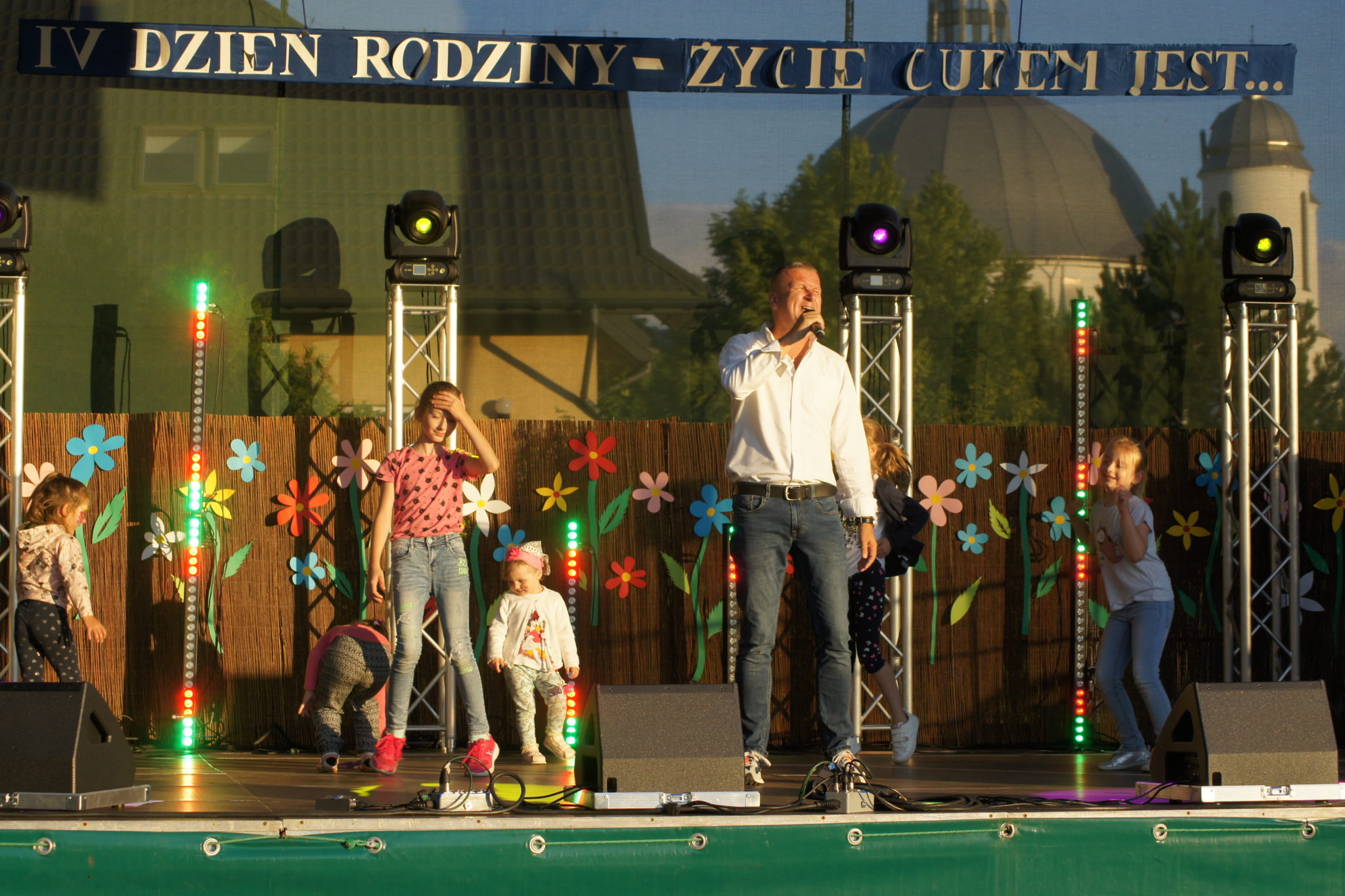 Mężczyzna śpiewający do mikrofonu i dzieci na scenie podczas IV Dnia Rodziny.