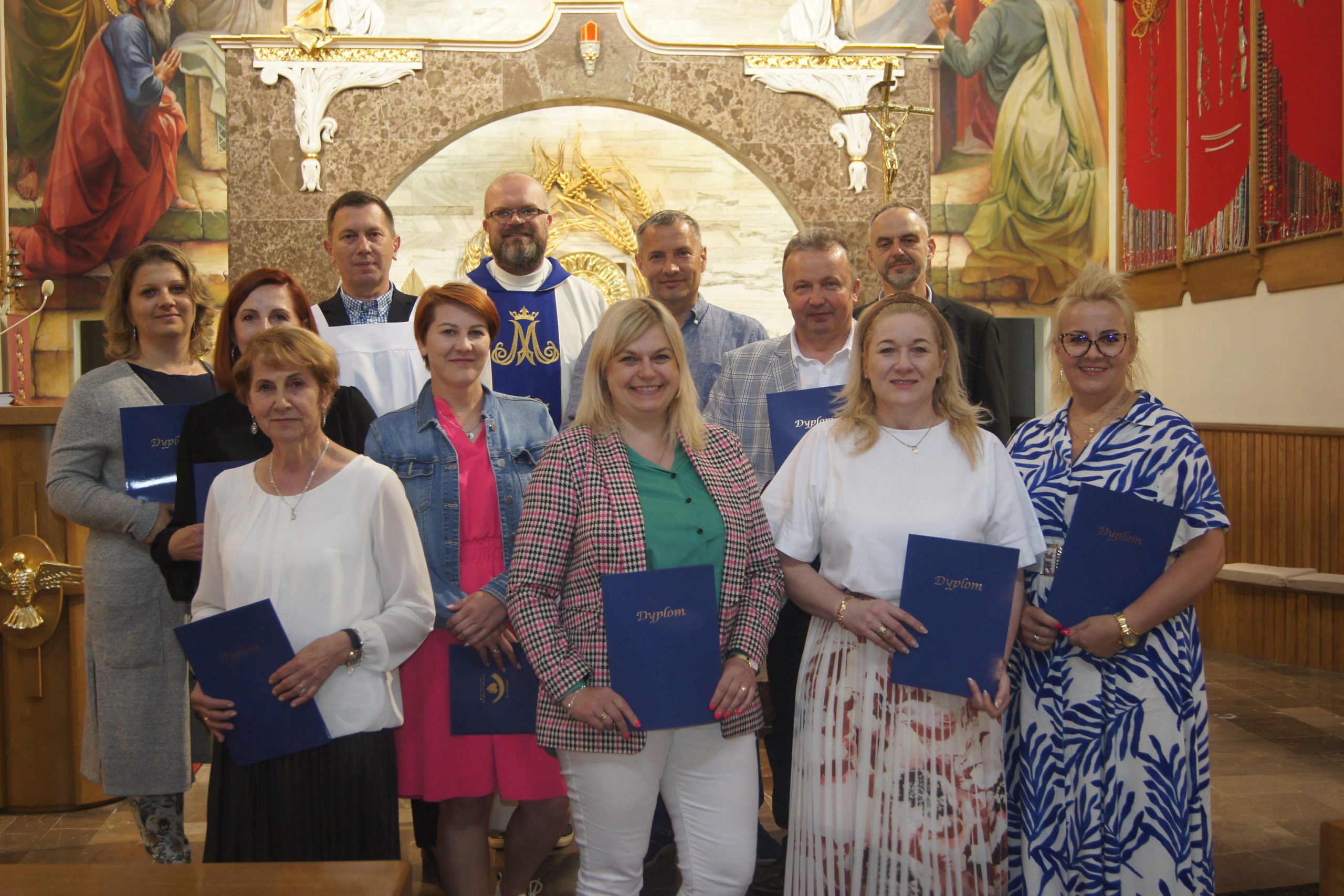 Grupa osób w kościele z dyplomami, w tle obraz religijny.