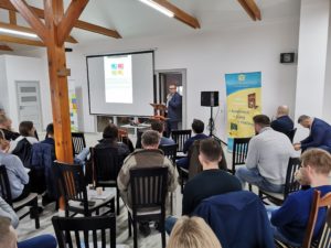 Prelegent przemawia do uczestników konferencji „Biblia o finansach”, stojąc przy mównicy obok ekranu projekcyjnego.