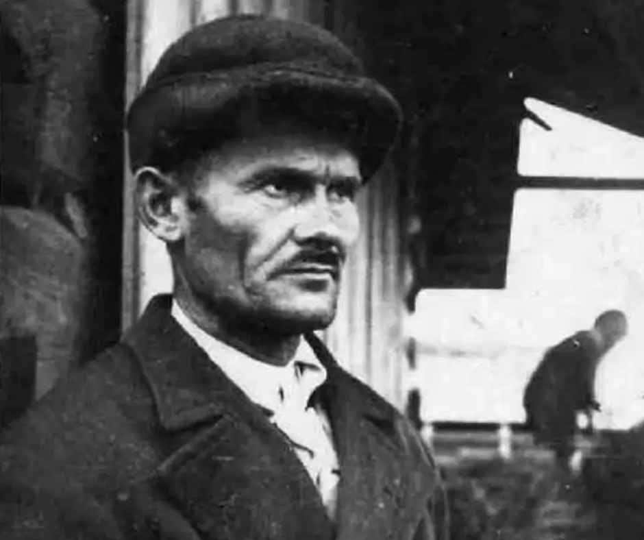 Zdjęcie mężczyzny w kapeluszu i płaszczu, stojącego zgarbionego. W tle widoczny jest inny człowiek.