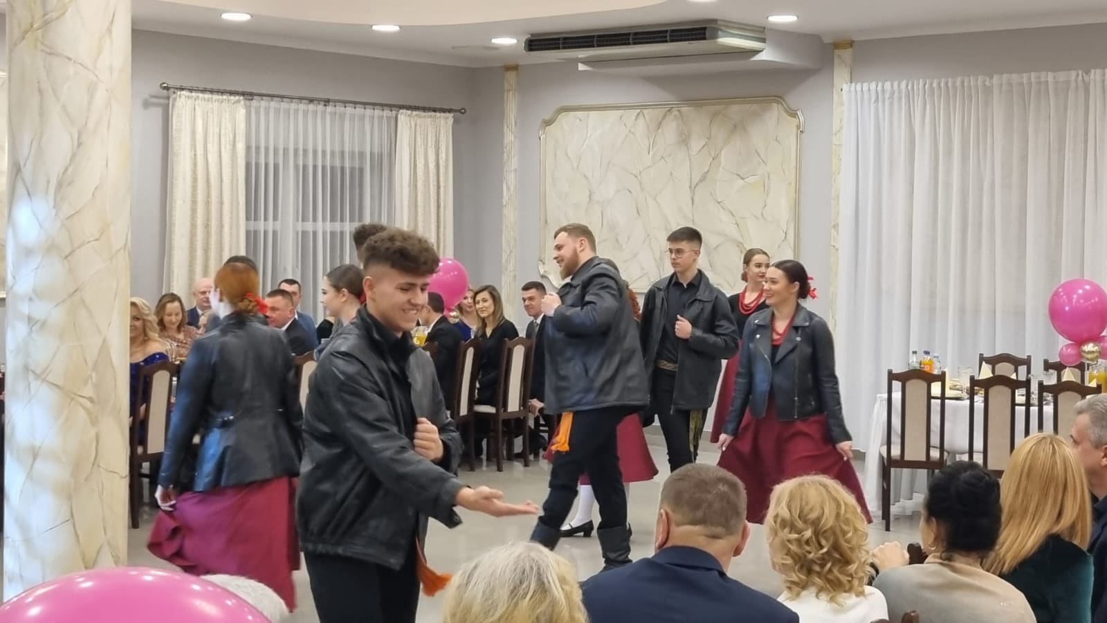 Uczestnicy balu tańczą w sali podczas wydarzenia charytatywnego. Widać mężczyznę i kobietę w trakcie tańca, a także inne osoby obserwujące. Na stole znajdują się różowe balony.