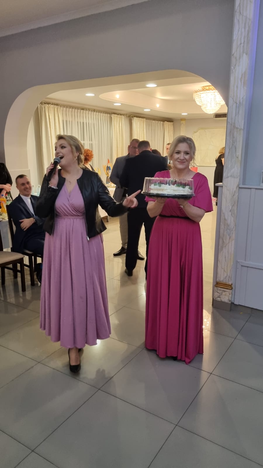 Na zdjęciu widać dwie osoby stojące w eleganckiej sali. Jedna z nich, kobieta w różowej sukni maxi, śpiewa do mikrofonu. Druga, kobieta w różowej sukience maxi, trzyma tort na talerzu. W tle widoczny mężczyzna i kilka innych osób.