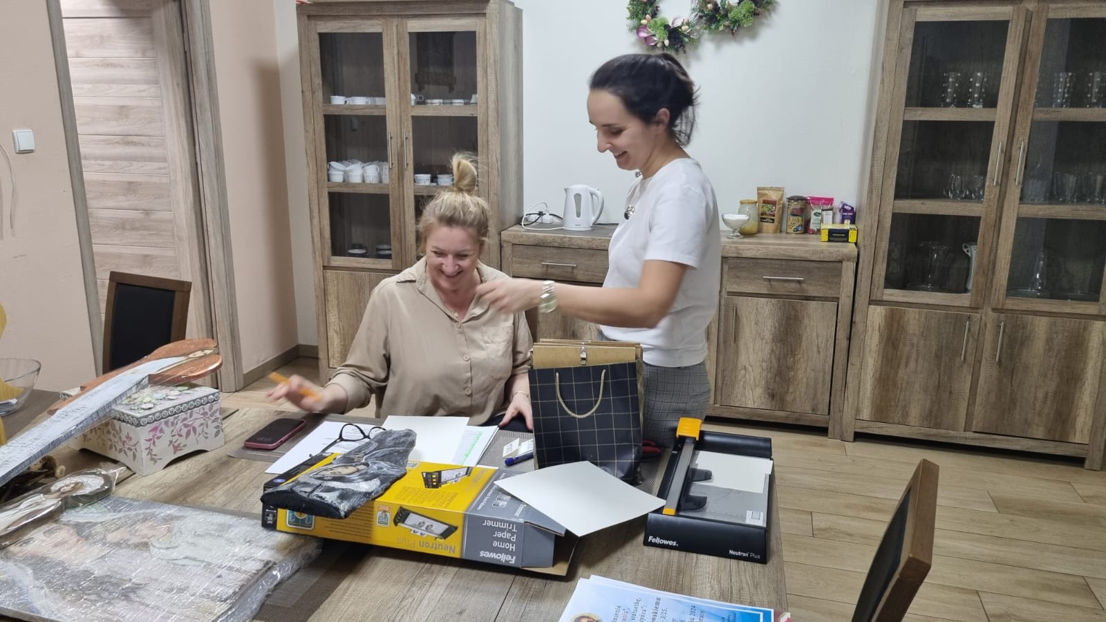 Dwie osoby składają elementy produktu na stole w pomieszczeniu. Na stole leżą instrukcje i materiały do montażu. W tle znajduje się szafka z szufladami.