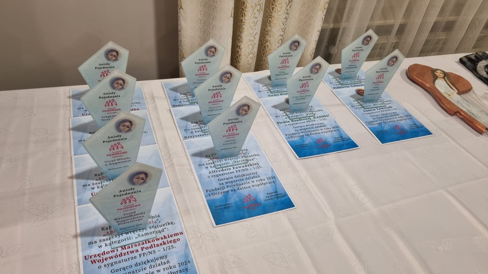 Na stole rozłożone są plakaty i ulotki z nagrodami Anioły Pojednania, przyznanymi podczas wydarzenia organizowanego przez Urząd Marszałkowski Województwa Podlaskiego. Plakaty zawierają zdjęcia osób oraz informacje o Fundacji Pojednanie.