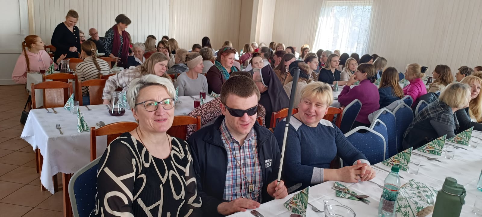 Zdjęcie przedstawia grupę osób podczas spotkania, siedzących przy stołach w sali restauracyjnej. Obywatele w różnym wieku i strojach ubrani w różne stroje. Na stole znajdują się napoje i dekoracje.