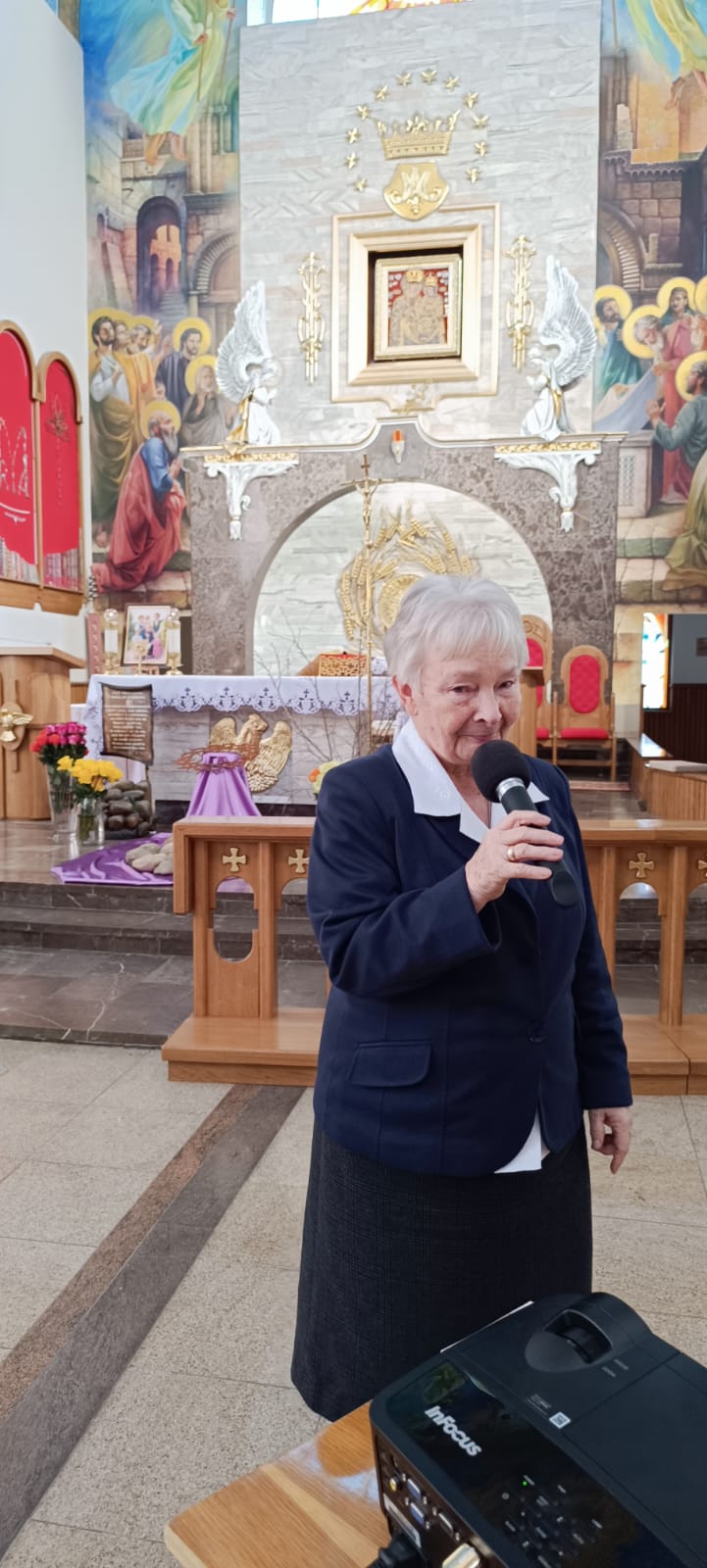 Kobieta trzyma mikrofon i przemawia podczas wydarzenia wewnątrz kościoła, na tle ołtarza z malowidłami oraz projektora InFocus. Widać elementy architektoniczne kościelne.