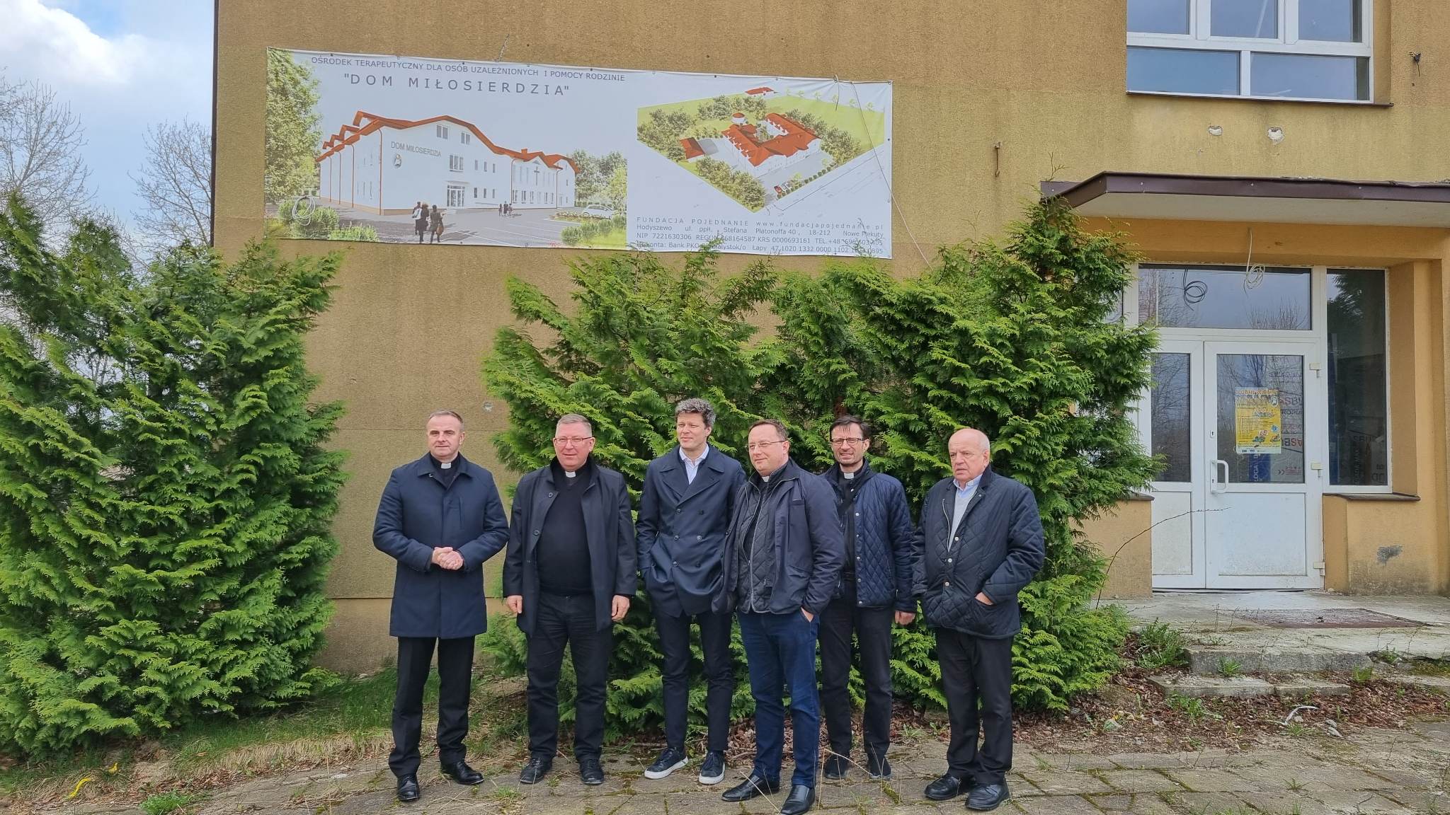 Grupa osób stoi przed budynkiem z plakatem. Plakat przedstawia rysunek budynku i teren.