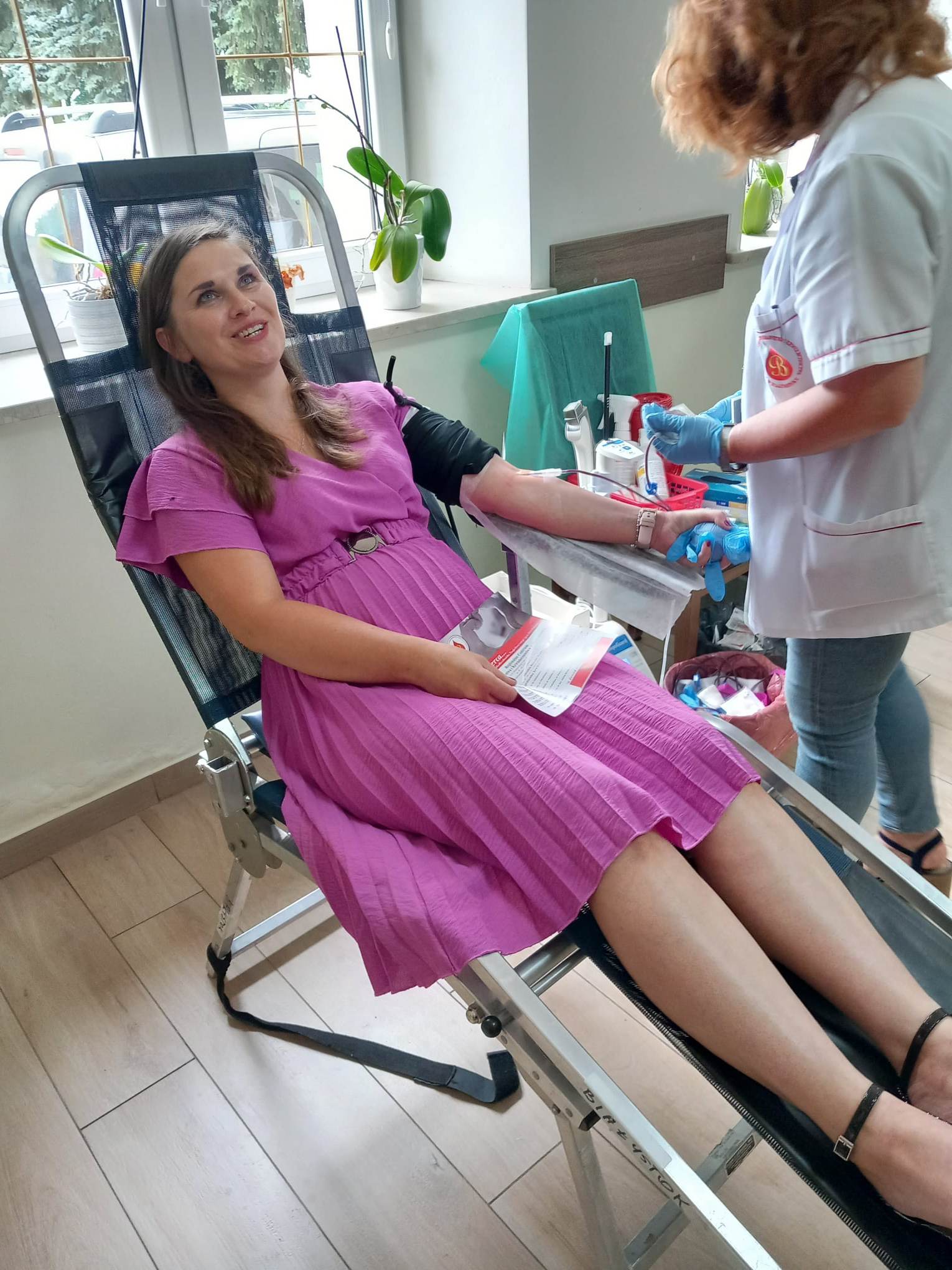 Kobieta siedzi na krześle do pobierania krwi, podczas akcji oddawania krwi. W tle personel medyczny przygotowuje sprzęt. Akcja odbywa się w centrum medycznym.