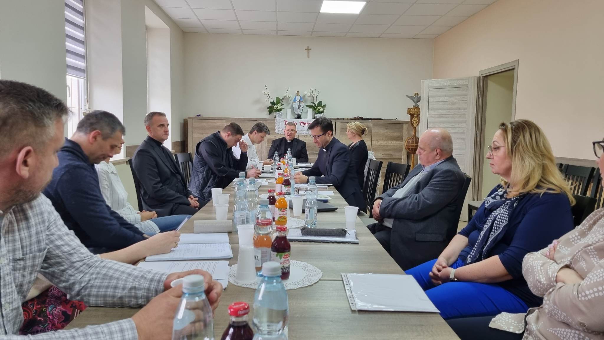 Grupa osób siedzi wokół stołu podczas spotkania. Na stole znajdują się dokumenty i napoje.