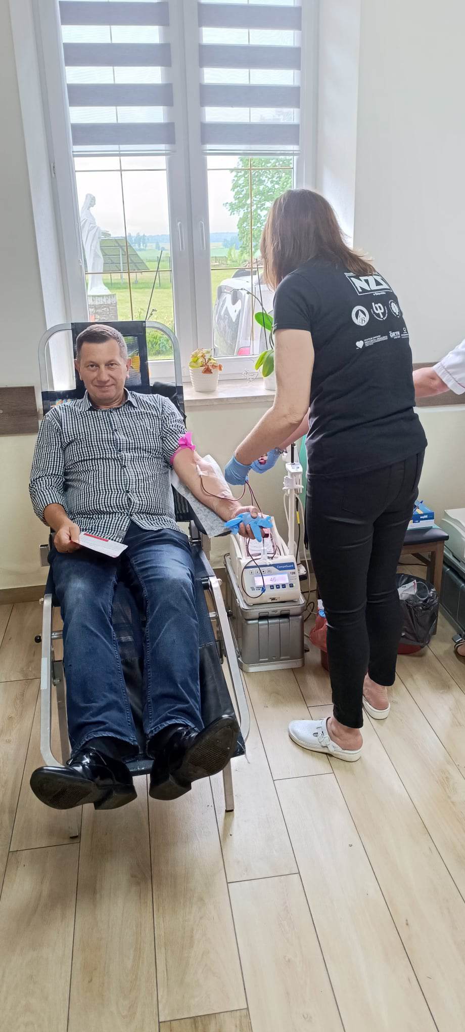 Osoba siedzi w fotelu do pobierania krwi, podczas gdy kolejna osoba wykonuje procedurę medyczną. W tle widoczne okno i pomieszczenie z podłogą z desek.